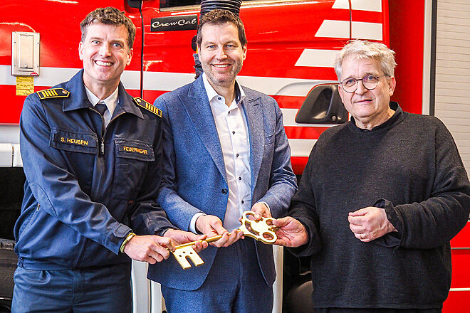 Drei Männer vor einem Feuerwehrauto halten einen großen goldenen Schlüssel.