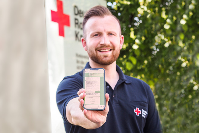 Mann mit DRK-Poloshirt zeigt das Display eines Handys mit einem WhatsApp-Chat-Verlauf.