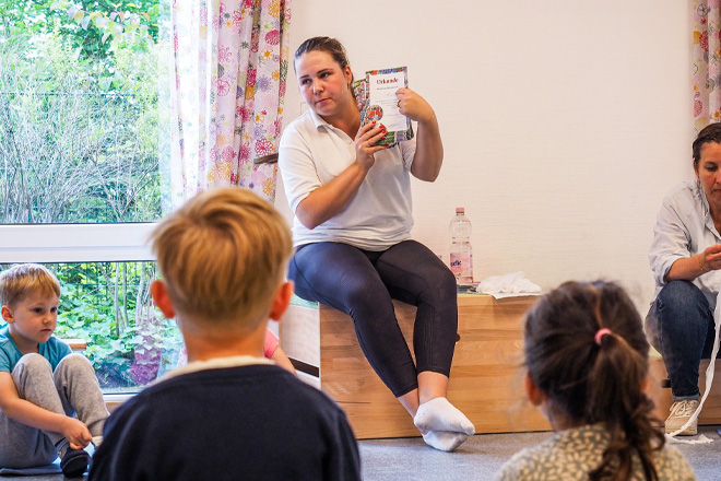 Frau auf einem Hocker hält eine Broschüre hoch; Kinder gucken zu.